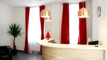 Ein Empfangsschalter mit Holzoptik, zwei Pendelleuchten, rote Vorhänge, eine kleine rote Weihnachtstanne und ein Topfpflanze.