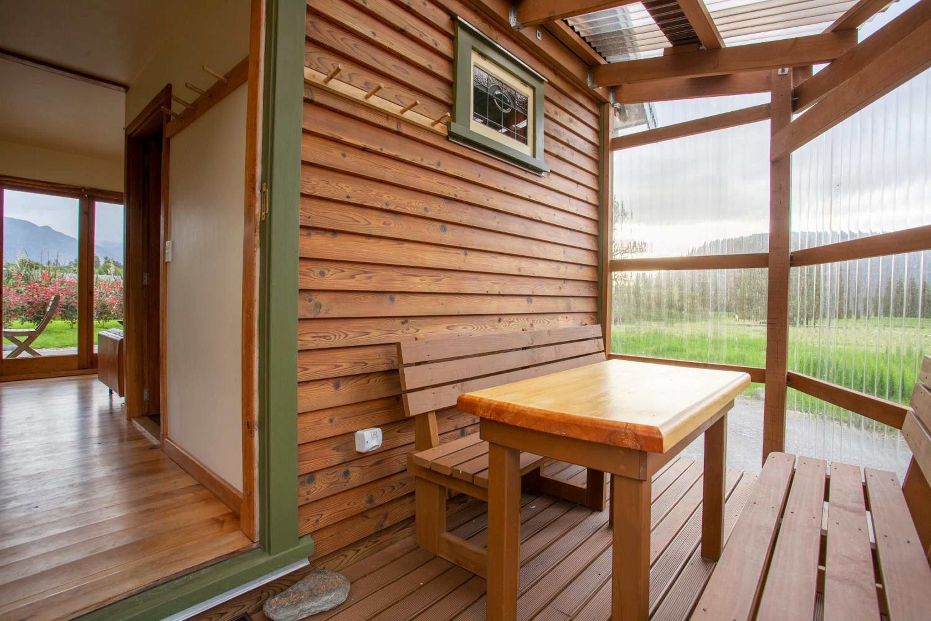 A wooden interior with a table and bench, a framed picture on the wall, and a view of greenery outside.