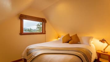 Quarto com cama, travesseiros, mesa de cabeceira, luminária e janela com vista para árvores.