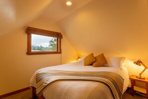 Un dormitorio con cama, almohadas, mesita de noche, lámpara y ventana con vistas a árboles.