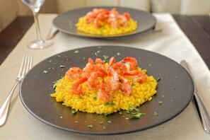 Dos platos de risotto amarillo con camarones sobre un plato oscuro, colocados sobre una mesa con un tenedor y una copa.