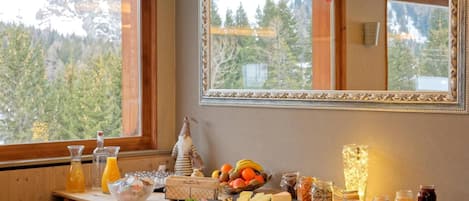 Un bufé de desayuno con diversos alimentos y bebidas, un espejo y vistas a árboles por la ventana.