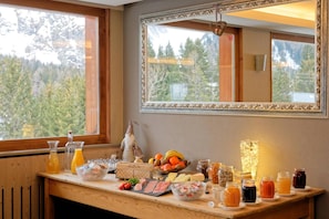 Un bufé de desayuno con diversos alimentos y bebidas, un espejo y vistas a árboles por la ventana.