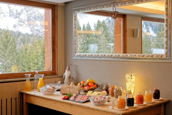 Un bufé de desayuno con diversos alimentos y bebidas, un espejo y vistas a árboles por la ventana.