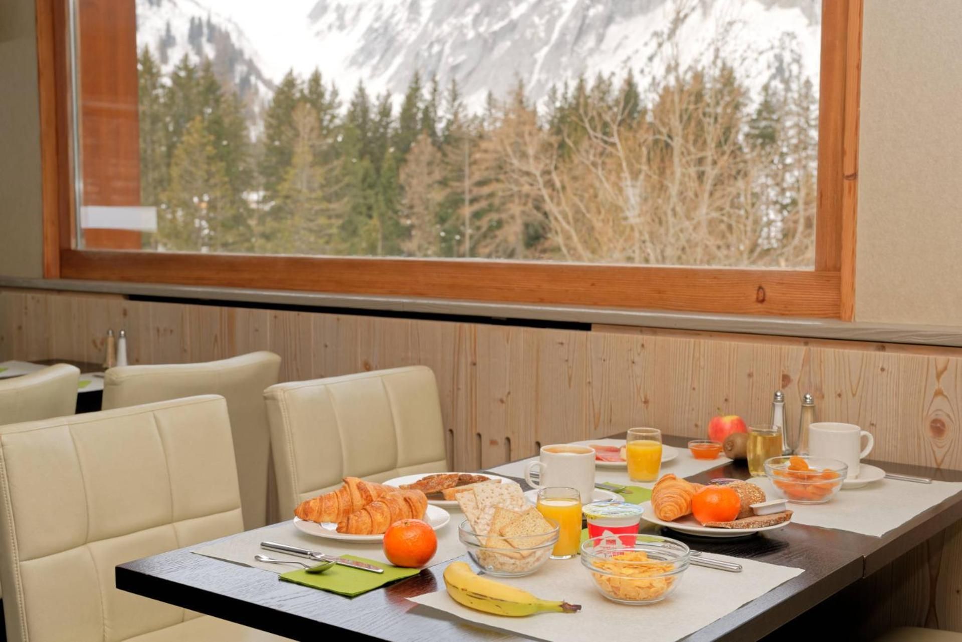 A breakfast table with croissants, fruits, and beverages, set against a backdrop of a forest and mountains.