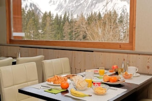 A breakfast table with croissants, fruits, and beverages, set against a backdrop of a forest and mountains.