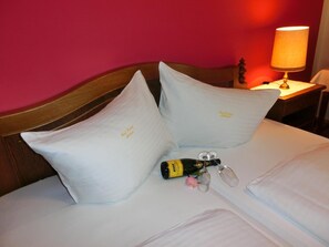 A hotel room with a wooden headboard, two white pillows, a bottle of wine, a glass, and a lamp on a bedside table. - Hotel Posthof (Millstatt)