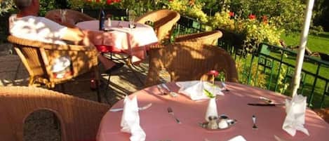 Un espace de restauration en plein air, avec des chaises et des tables en osier disposées pour un repas, entouré de verdure et avec un bâtiment en arrière-plan.