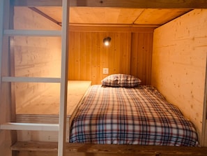 Una cama individual con colcha a cuadros en una habitación tipo cabaña, con paredes de madera y techo inclinado.