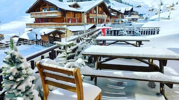 A snow-covered outdoor seating area with wooden benches and a chair, surrounded by buildings with wooden facades and snow-covered roofs.
