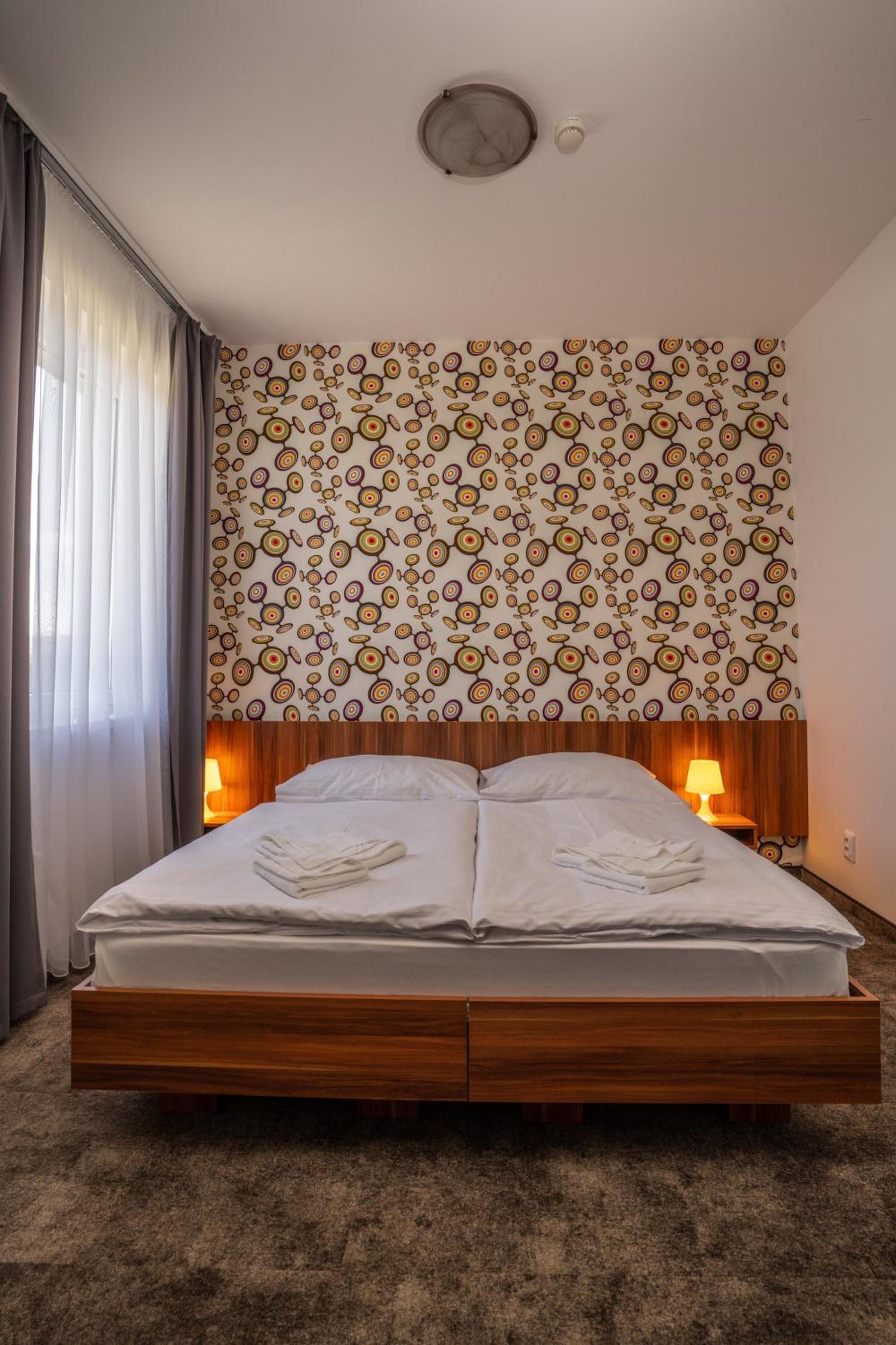 A hotel room with a wooden bed frame, a patterned wall, and two bedside lamps.
