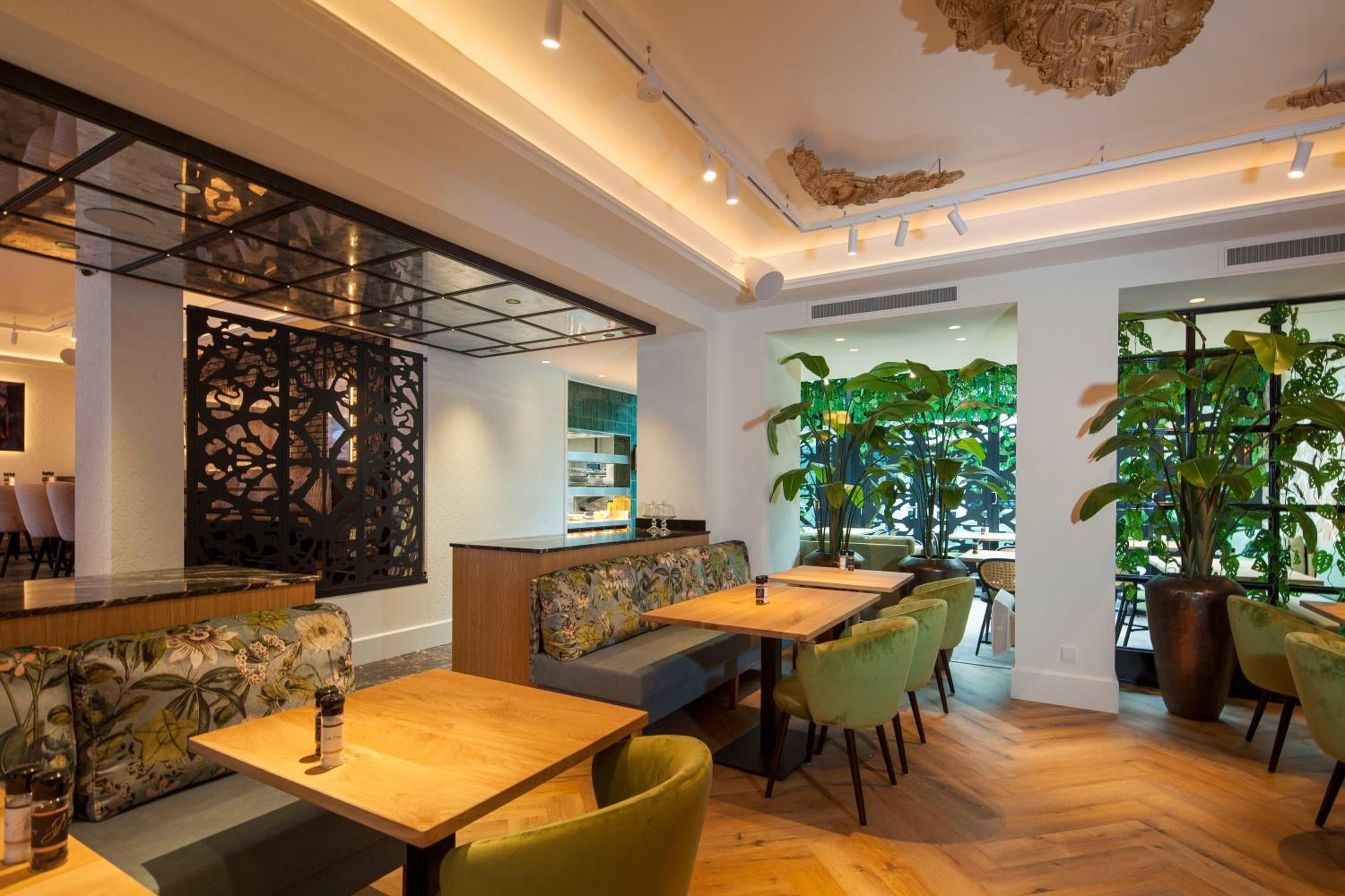 Een modern interieur met houten tafels, beklede zitplaatsen in groen en grote ramen met binnenplanten. Een decoratief plafond completeert het geheel.