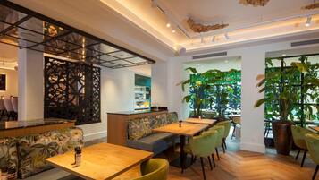 Een modern interieur met houten tafels, beklede zitplaatsen in groen en grote ramen met binnenplanten. Een decoratief plafond completeert het geheel.