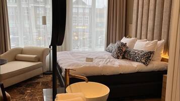 A hotel room with a bed, a sofa, a bedside table with a bowl, and a view of buildings through the window.