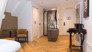 A hotel room with a wooden herringbone floor, a bed, a chair, a small table, a desk with a computer, and a bathroom with a glass shower enclosure.