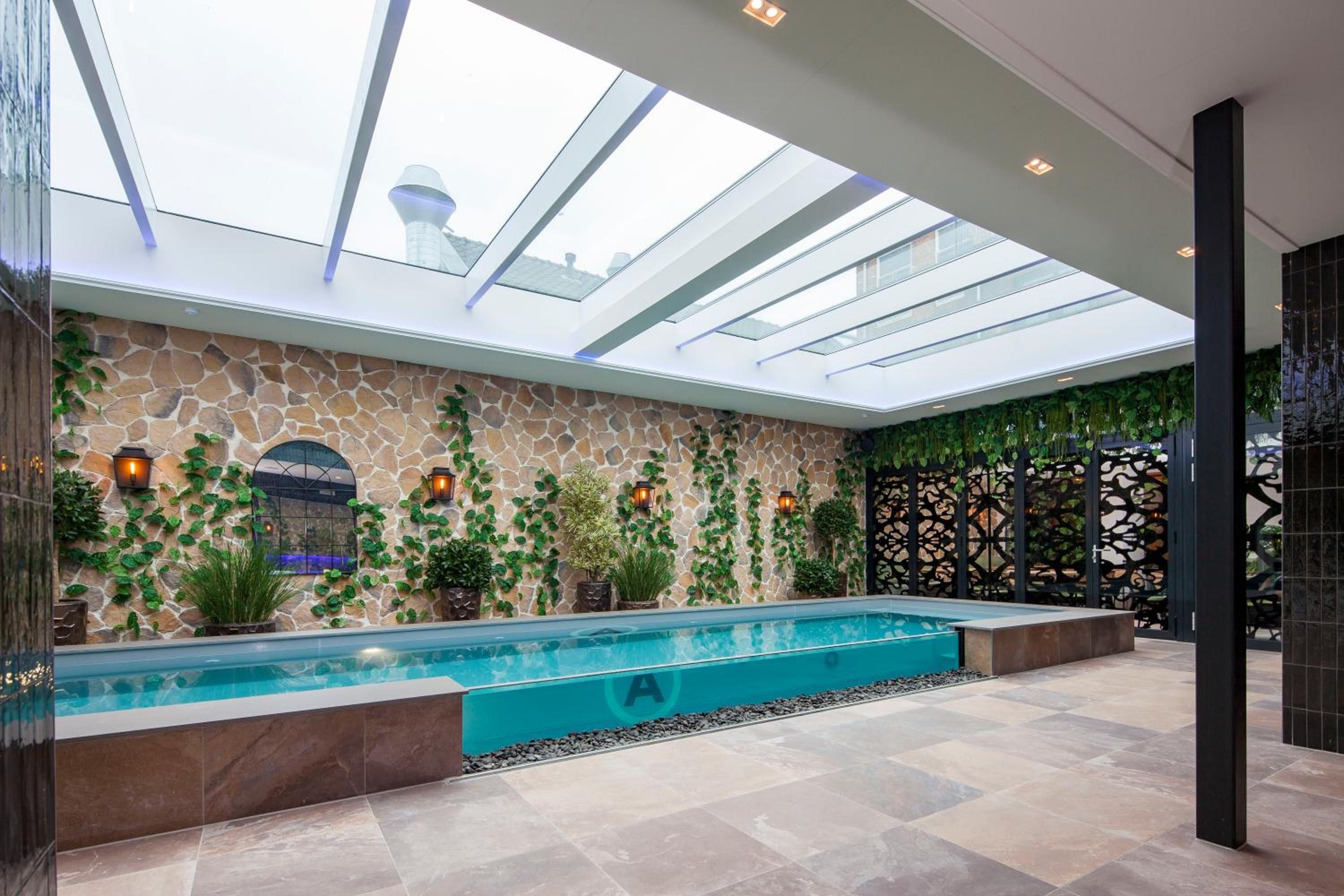 Área interna de piscina moderna, com claraboia, paredes de pedra e plantas em vasos.