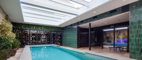 A modern indoor pool area with a skylight ceiling, a pool with steps leading into it, and a glass door leading to an outdoor area.