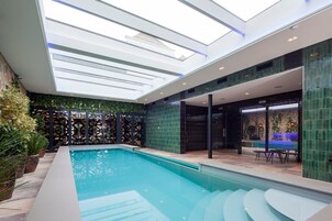 Un área de piscina interior moderna con techo de tragaluces, una piscina con escalones de acceso y una puerta de cristal que da a un espacio exterior.