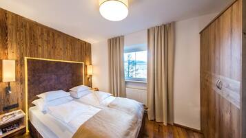 Ein Hotelzimmer mit Holz-Kopfteil, einem großen Bett mit weißer Bettwäsche, einem Nachttisch mit Lampe, einem Fenster mit Vorhängen und einem Kleiderschrank.