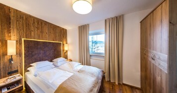 A hotel room with a wooden headboard, a large bed with white linens, a bedside table with a lamp, a window with curtains, and a wardrobe.