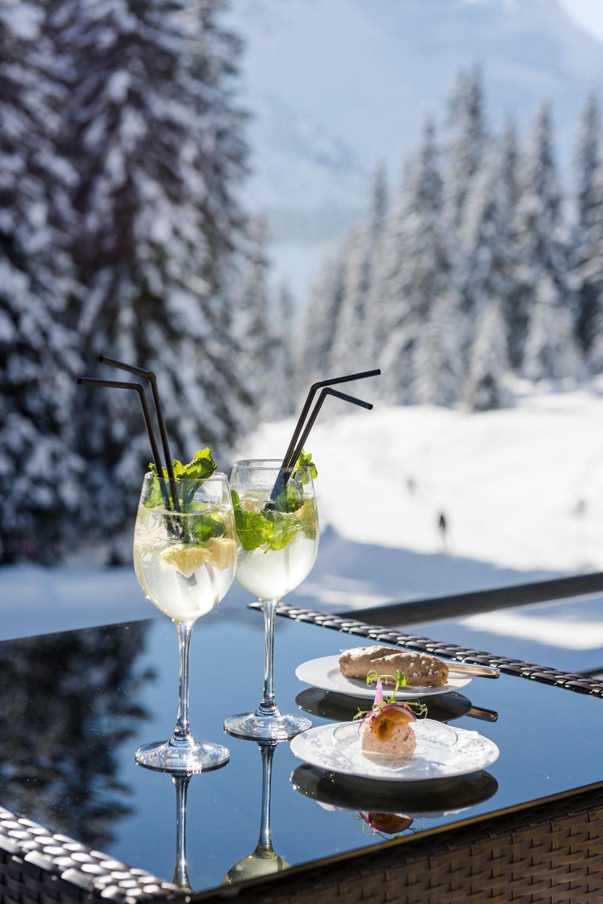 Two glasses of a clear beverage with mint leaves and straws, and two plates with food items on a reflective surface with a snowy mountain background.