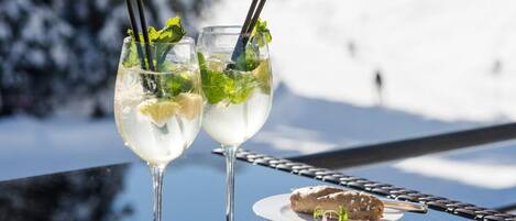 Two glasses of a clear beverage with mint leaves and straws, and two plates with food items on a reflective surface with a snowy mountain background.