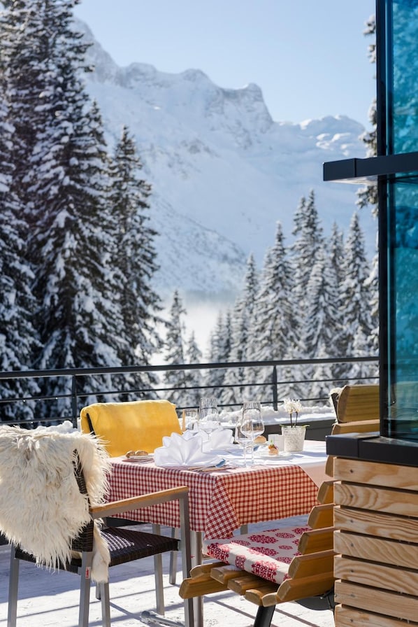 Un área de comedor al aire libre cubierta de nieve con una mesa lista para dos, con vista a un paisaje montañoso.