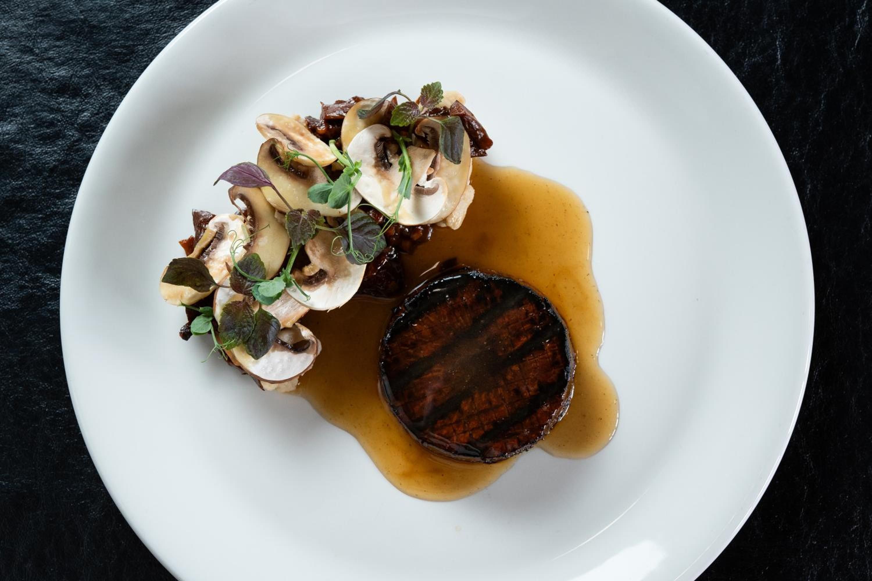 A plate with a dark, round food item and sliced mushrooms with greens on a white plate.