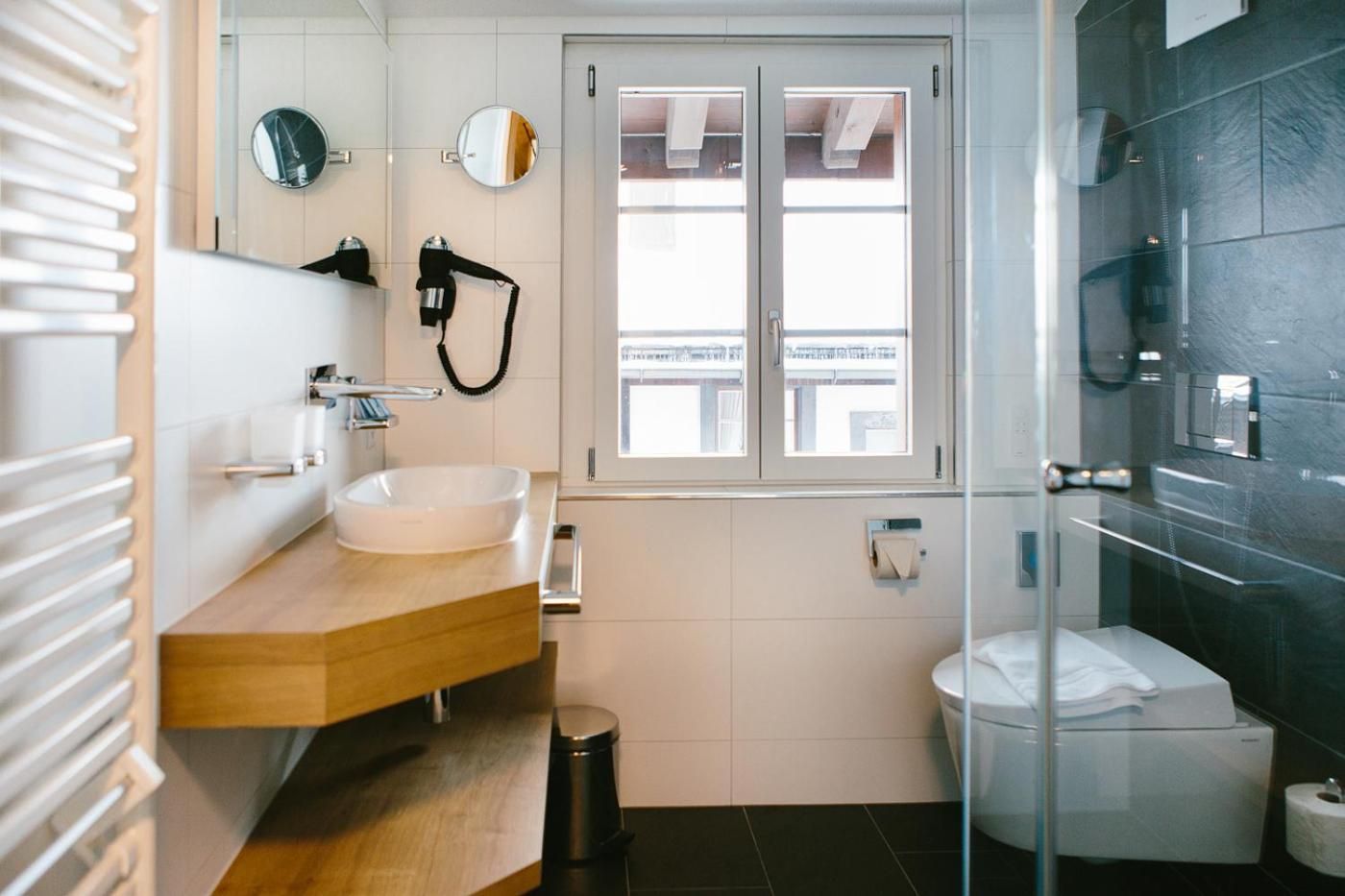 Ein modernes Badezimmer mit Waschtisch aus Holz, einem weißen Waschbecken, einer Duschkabine aus Glas und einem Fenster für natürliches Licht.