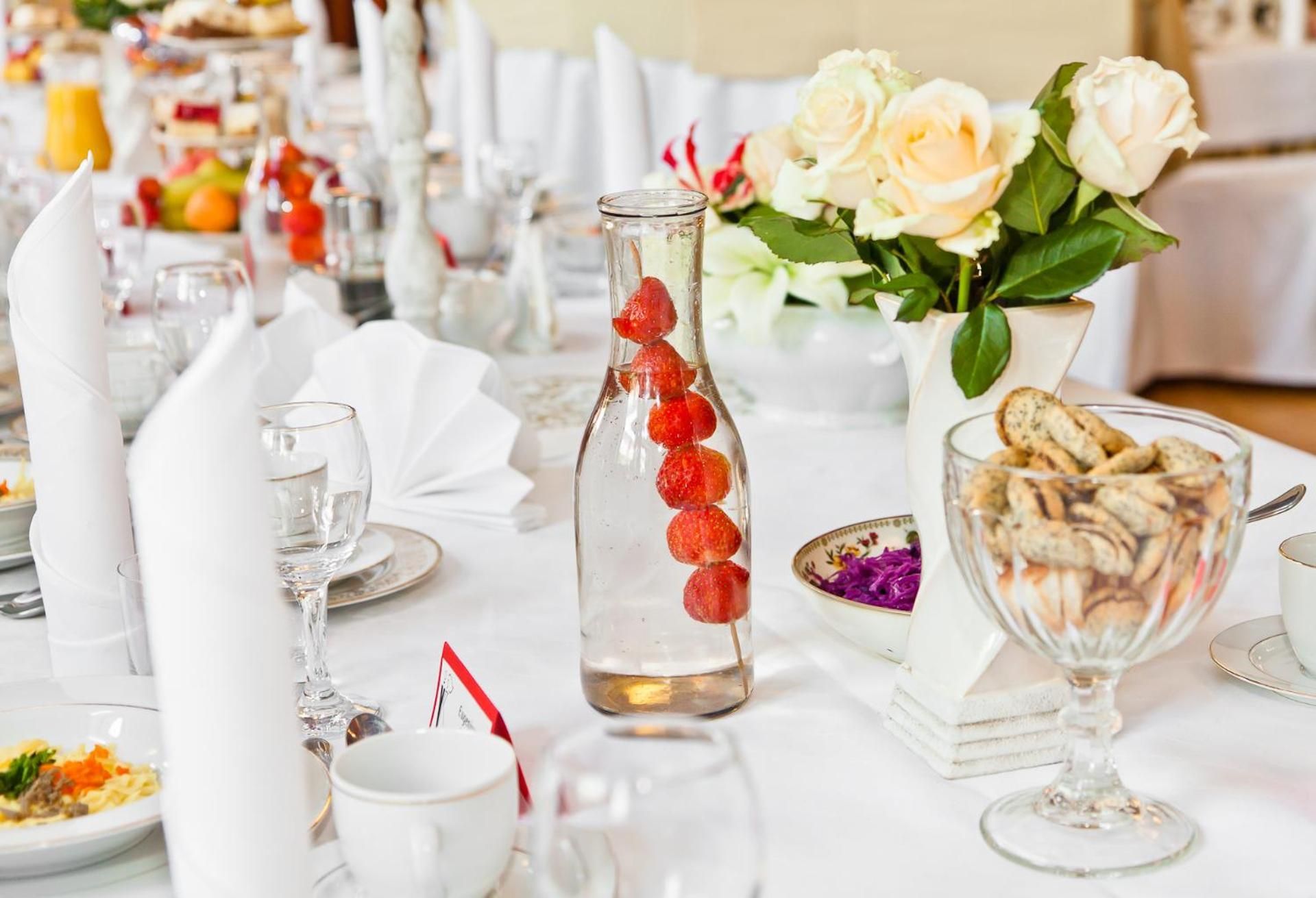 Een gedekte tafel met witte servetten, glazen, een fles met rode elementen, een vaas met witte bloemen en een schaal met lekkernijen.