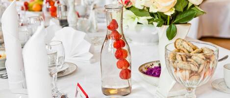 A table set with white napkins, clear glasses, a glass bottle with red objects, a vase with white flowers, and a bowl of snacks.