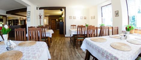 Un espace repas avec des tables et des chaises en bois, une cuisine à l’arrière-plan et une vue sur l’extérieur grâce à de grandes fenêtres.