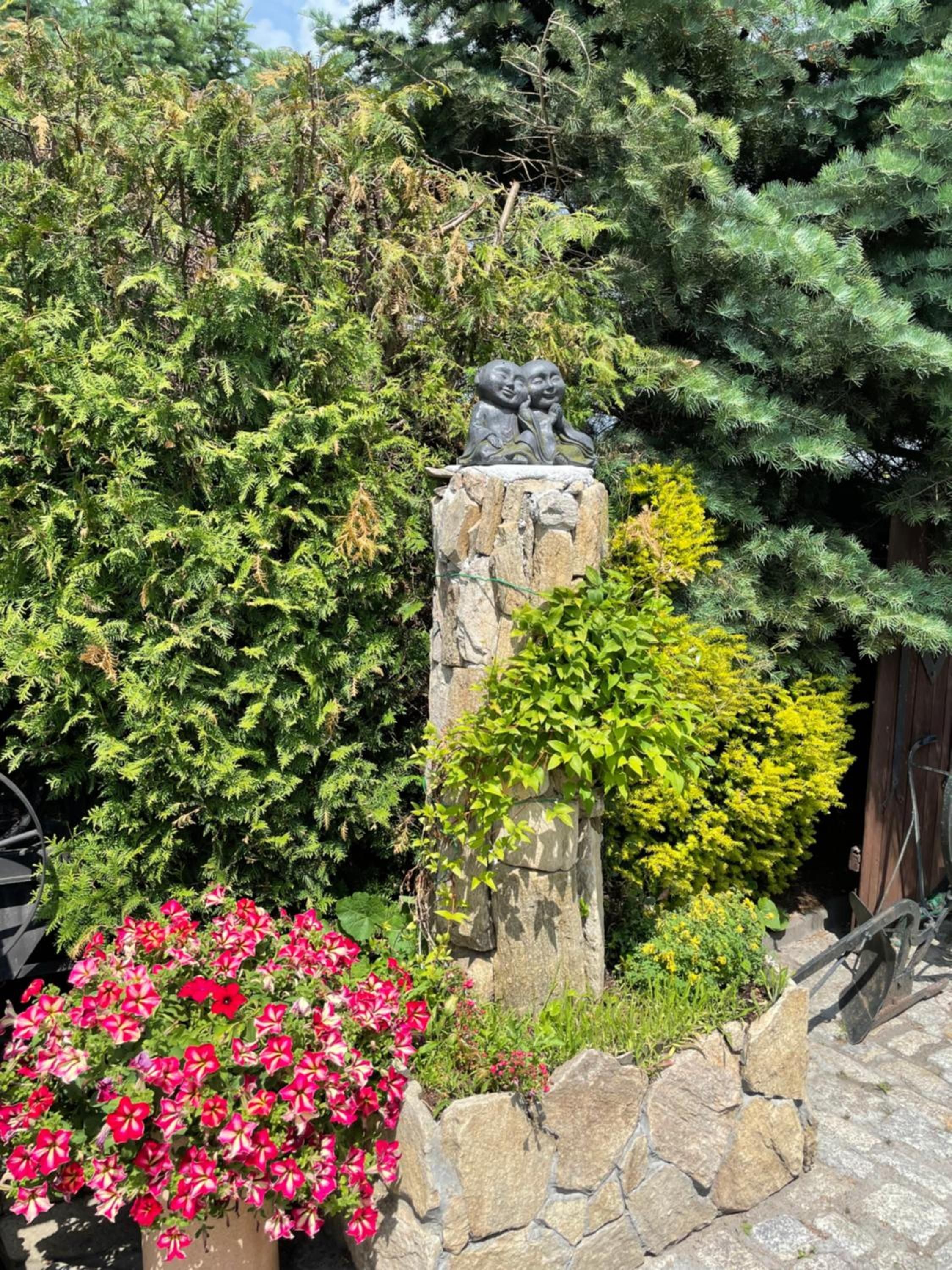 A stone pillar with a sculpture, surrounded by lush greenery and flowering plants.