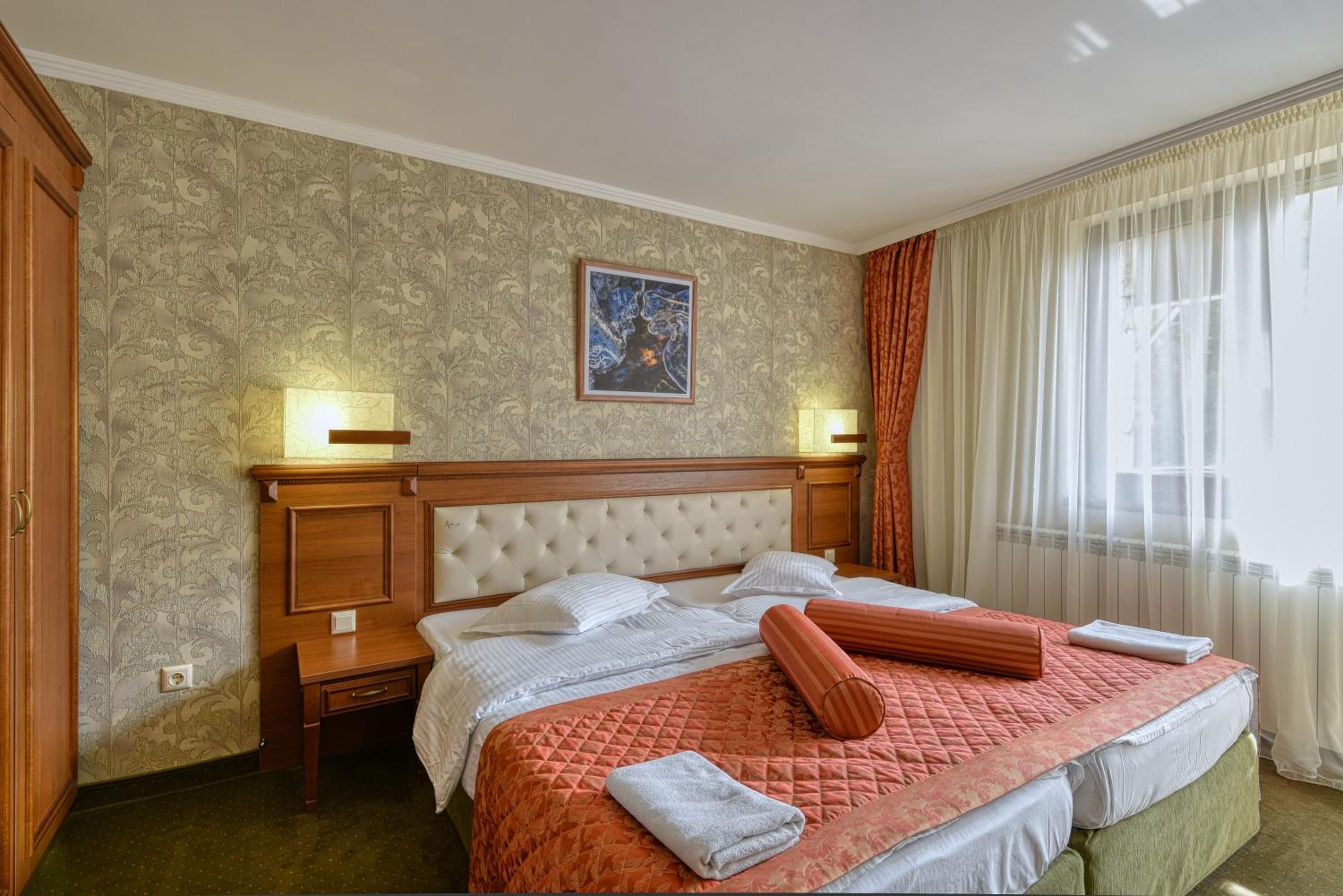 A hotel room with a large bed, two bedside tables, a nightstand, a painting on the wall, and a window with curtains.