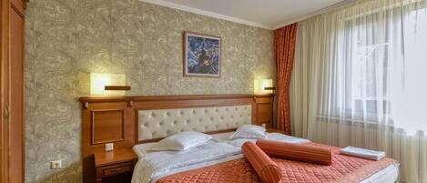 A hotel room with a large bed, two bedside tables, a nightstand, a painting on the wall, and a window with curtains.