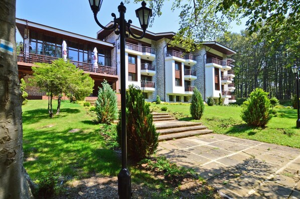 Um edifício de vários andares com varandas, fachada de pedra e um jardim bem cuidado com um caminho de pedra que leva à entrada.