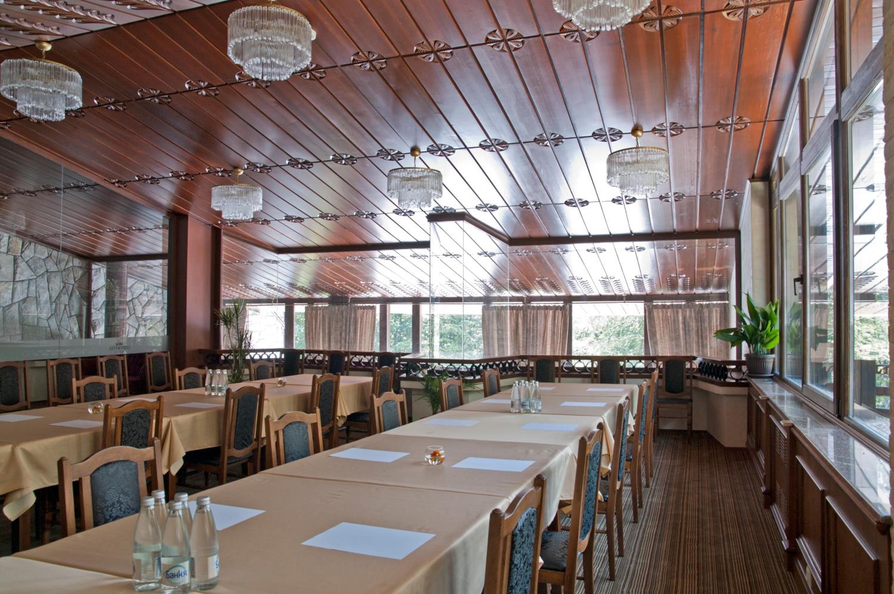 A restaurant with wooden panel ceilings, chandeliers, and large windows. Tables are set with white tablecloths and chairs.
