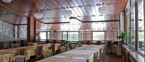 A restaurant with wooden panel ceilings, chandeliers, and large windows. Tables are set with white tablecloths and chairs.