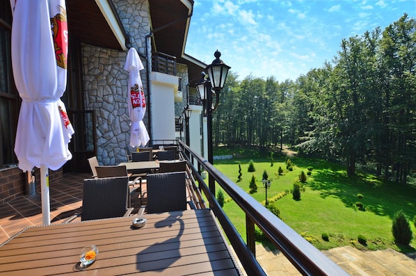 A wooden deck with outdoor furniture, a stone wall, and a view of a green landscape. - Hotel Edelweiss (Kazanlak)