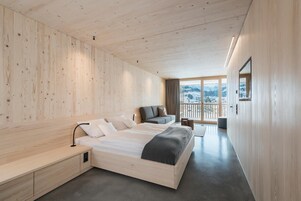 Quarto moderno com teto de madeira, cama grande, sofá e vista para montanhas pela janela.