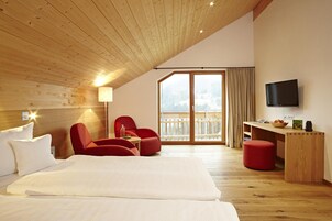 A hotel room with a wooden ceiling, a bed, two red chairs, a desk with a TV, and a balcony with a view.