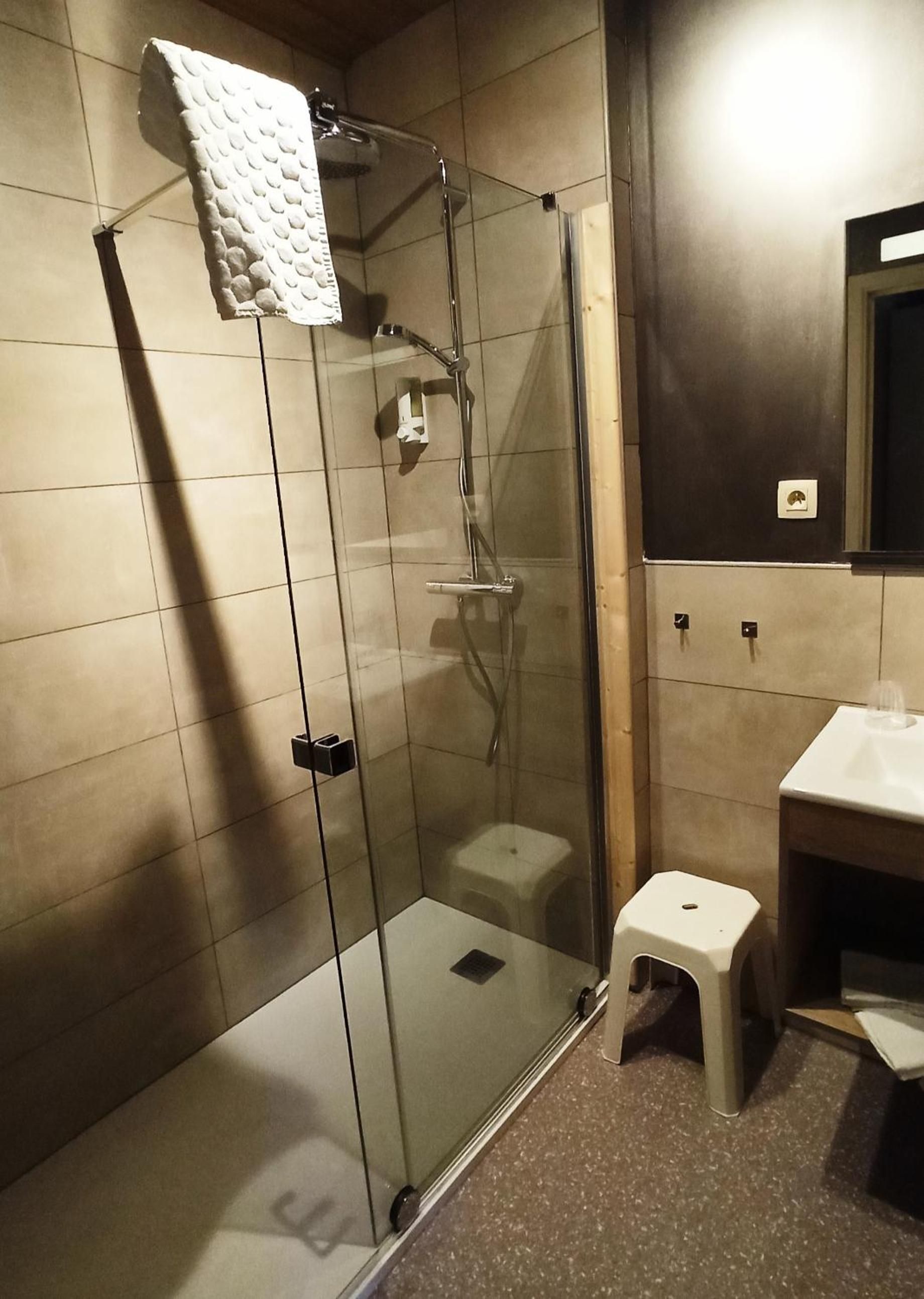 Een douchecabine met glazen wanden, een witte handdoek hangt aan de deur, een klein krukje en een wastafel met een spiegel erboven.