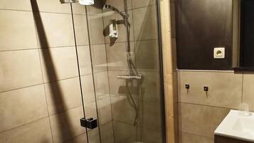 Een douchecabine met glazen wanden, een witte handdoek hangt aan de deur, een klein krukje en een wastafel met een spiegel erboven.