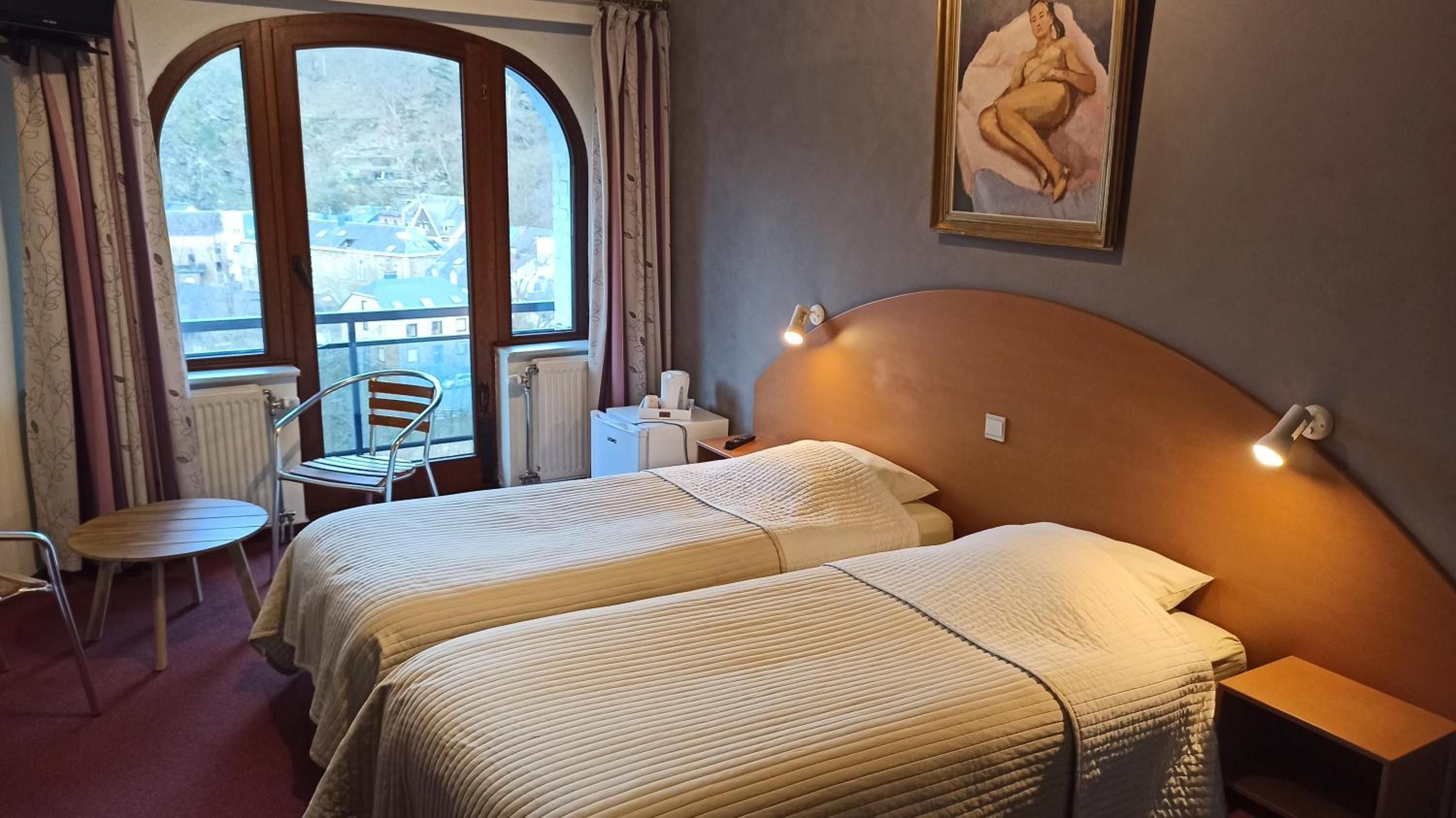 Een hotelkamer met twee bedden, een tafeltje, een stoel, een radiator en een raam met gordijnen.