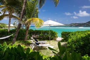 Un área de descanso junto a la playa con sillones blancos y sombrillas, rodeada de exuberante vegetación y con vistas a un mar azul cristalino.