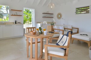 Cozinha moderna com armários brancos, sala de jantar com cadeiras de madeira e uma mesa redonda, e vista para palmeiras pela janela.