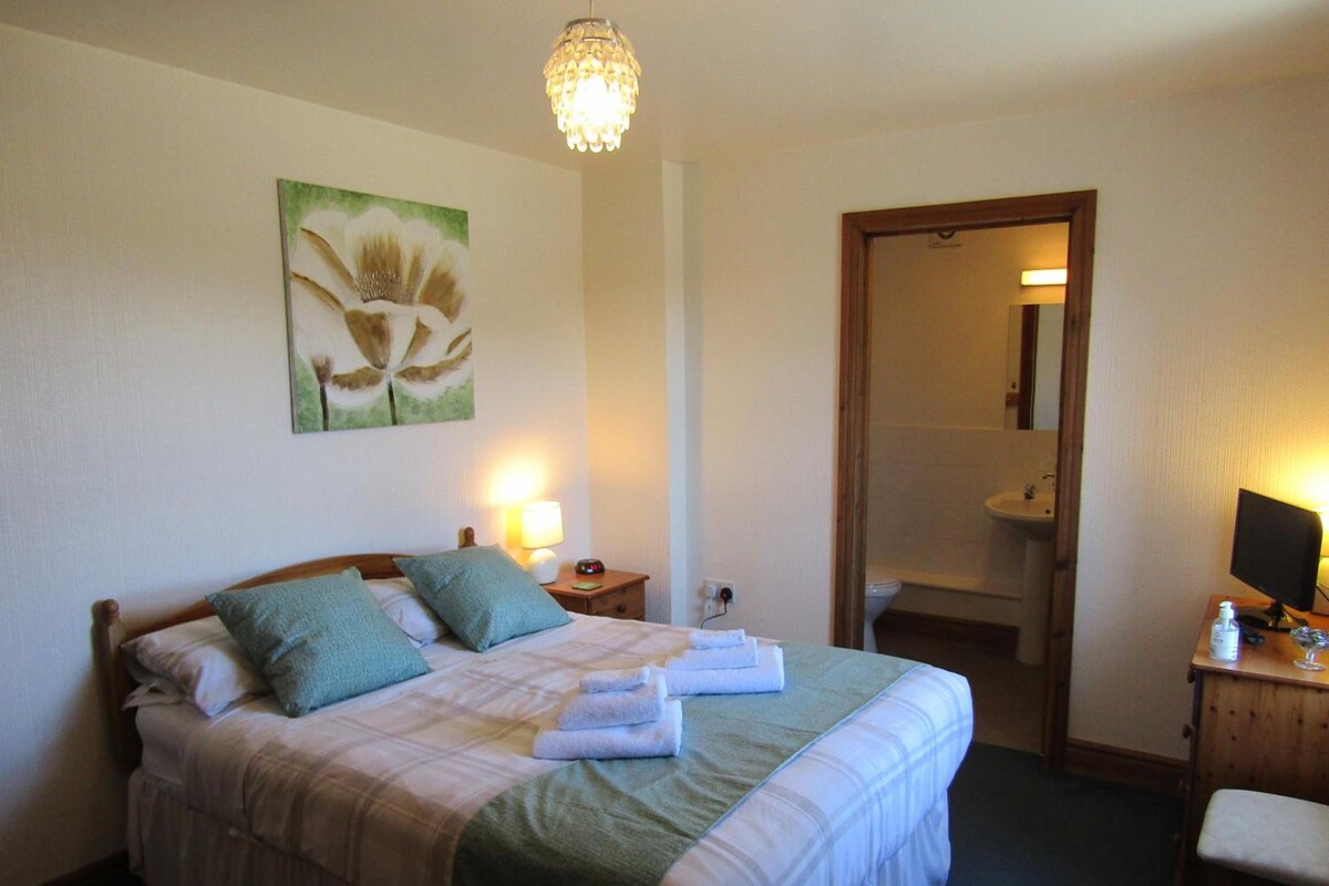 Une chambre d’hôtel avec un lit, une table de chevet avec une lampe, un tableau au mur et une salle de bain visible à travers une porte ouverte.