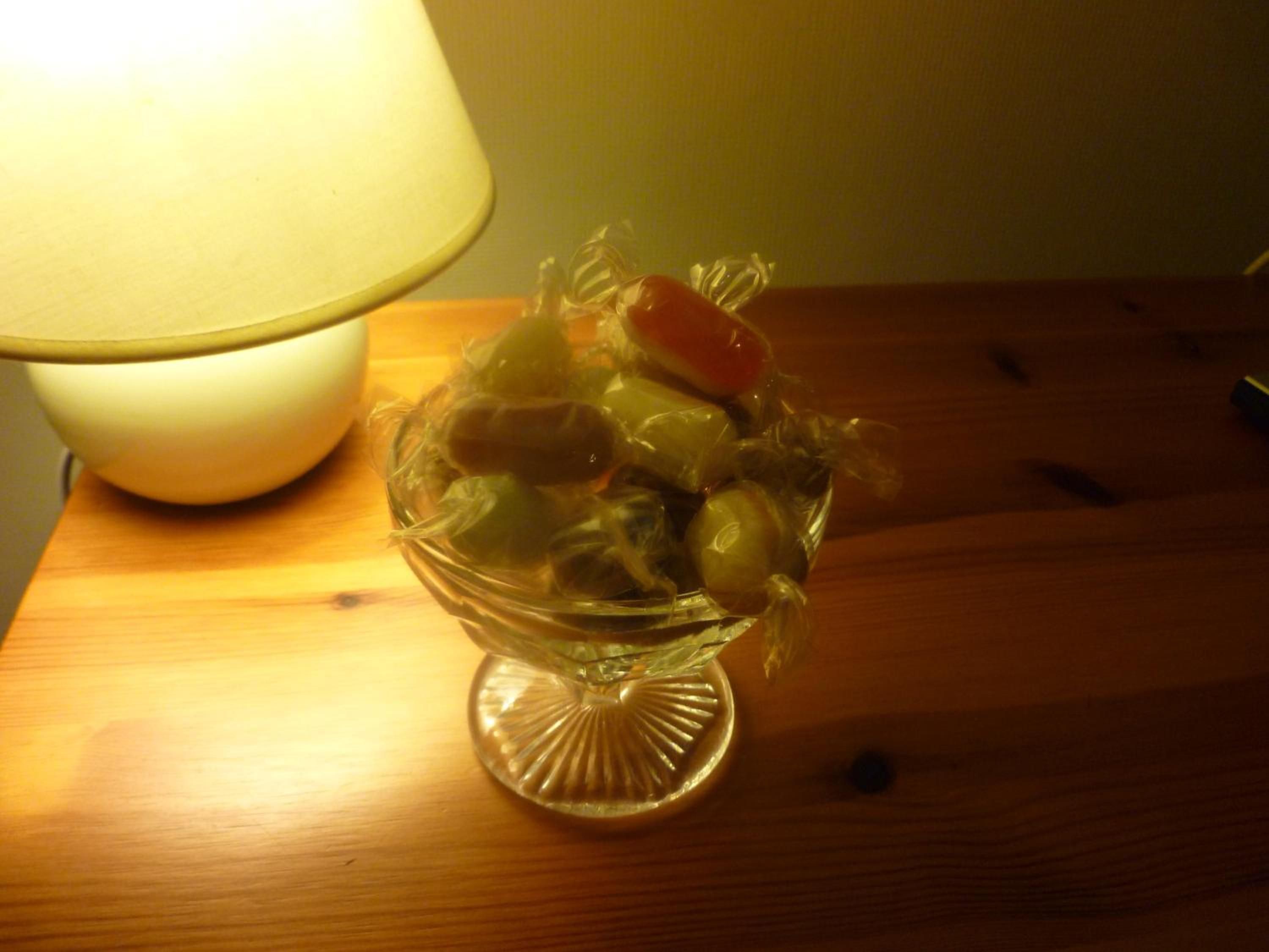 A clear glass bowl with various fruits on a wooden table beside a lamp.