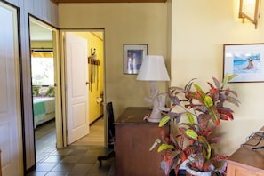 A hotel room with a bed, a desk with a lamp, a plant, and a door leading to another room.