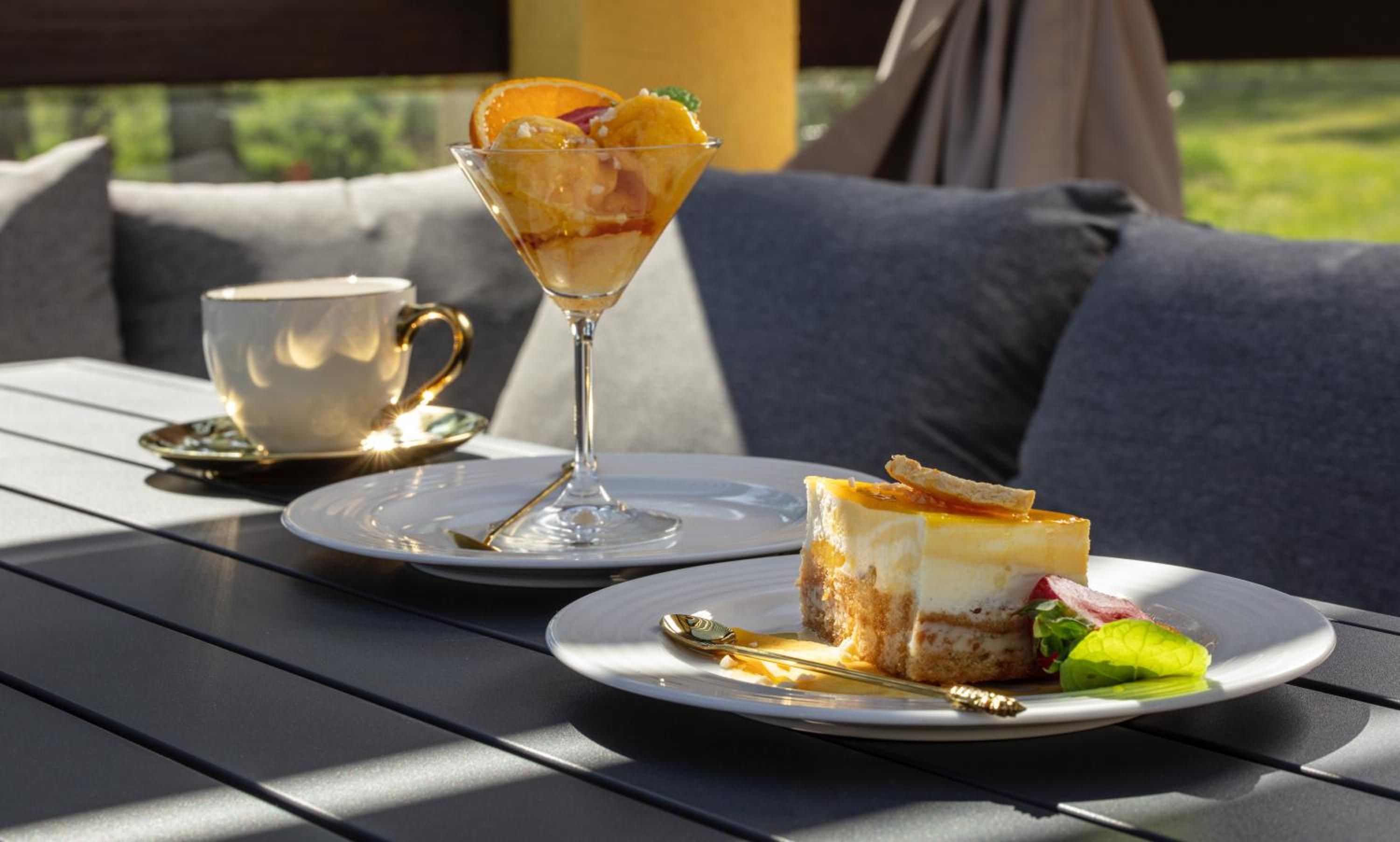 A table with a dessert, a glass beverage, and a cup, set on a sunny outdoor table.